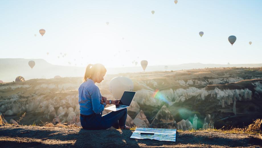 Back view of young woman sitting near map and using laptop while sitting against rocky terrain and colorful hot air balloons against cloudless sky in back lit