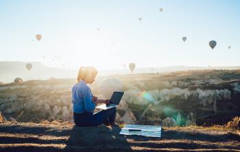 Back view of young woman sitting near map and using laptop while sitting against rocky terrain and colorful hot air balloons against cloudless sky in back lit
